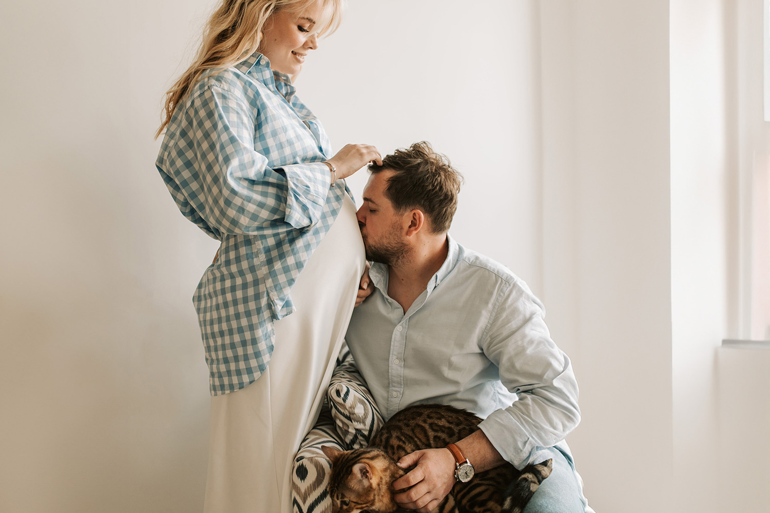 une femme enceinte se tient debout, son partenaire assis sur un fauteuil embrasse son ventre arrondi.