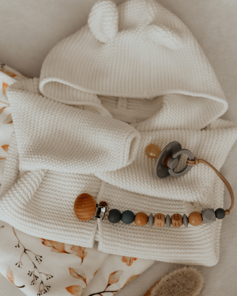 petite veste en laine à capuche pour bébé de couleur blanche, accompagnée d’une tétine avec son attache-tétine en bois