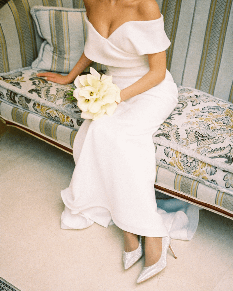 Une mariée assise sur un canapé, portant une longue robe sirène et des chaussures à bout pointues fermées de couleur blanche.