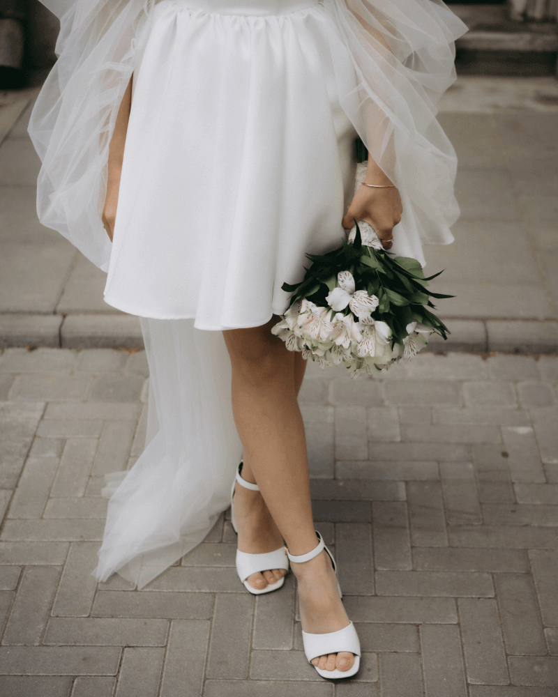 une mariée portant une robe courte au-dessus du genou et tenant un bouquet de fleurs blanches. À ses pieds, des sandales blanches ouvertes.