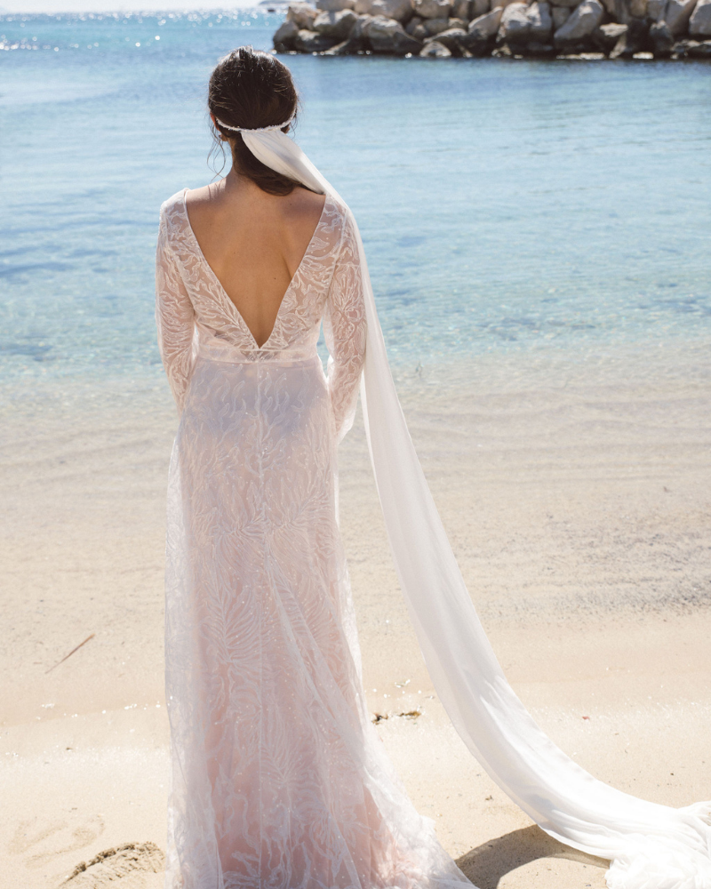 Une femme vue de dos, faisant face à la mer et portant une robe de mariée avec dos décolleté en V. Son long voile de mariée est attaché grâce à une couronne blanche.