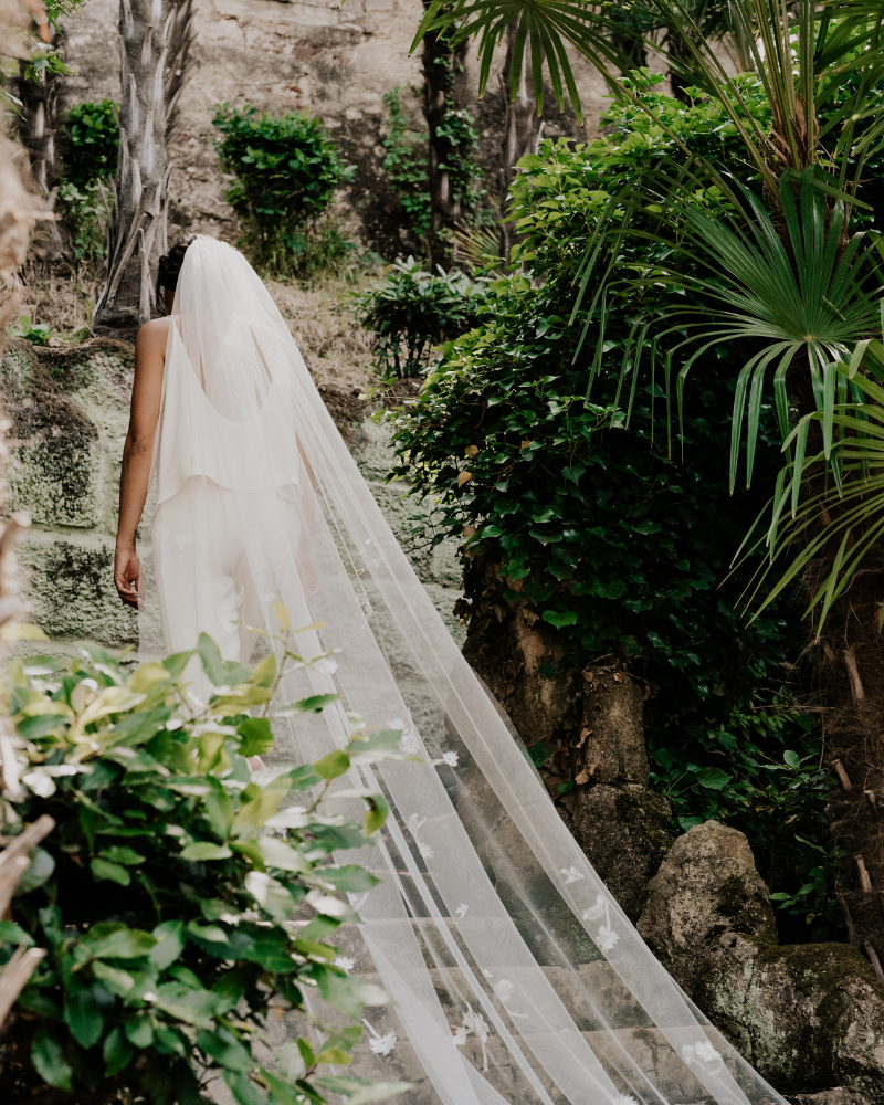 Une jeune mariée de dos, montant les escaliers à l'extérieur, dans un décor de jungle. Son voile cathédrale traîne derrière elle élégamment.