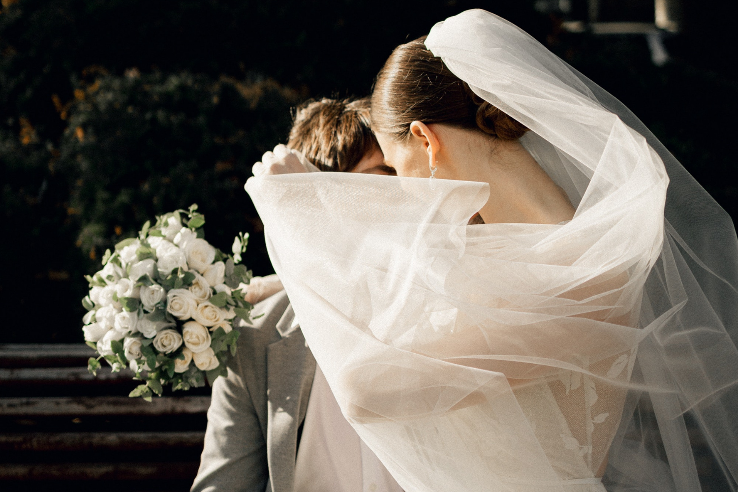 Un couple de jeunes mariés s'enlaçant, le voile de la mariée en premier plan cachant leurs visages.