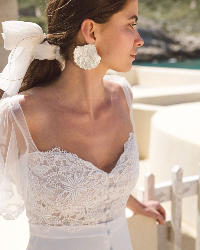 Une jeune mariée vue de profil portant une robe joliment brodée, des boucles d'oreilles coquillage et un voile de mariée noué dans ses cheveux.