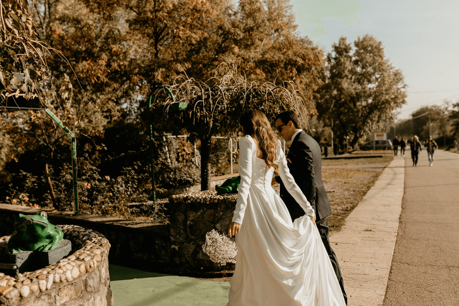 Un couple de jeunes mariés de dos, la mariée tenant un bouquet de fleurs blanches dans une main et sa robe dans l’autre.