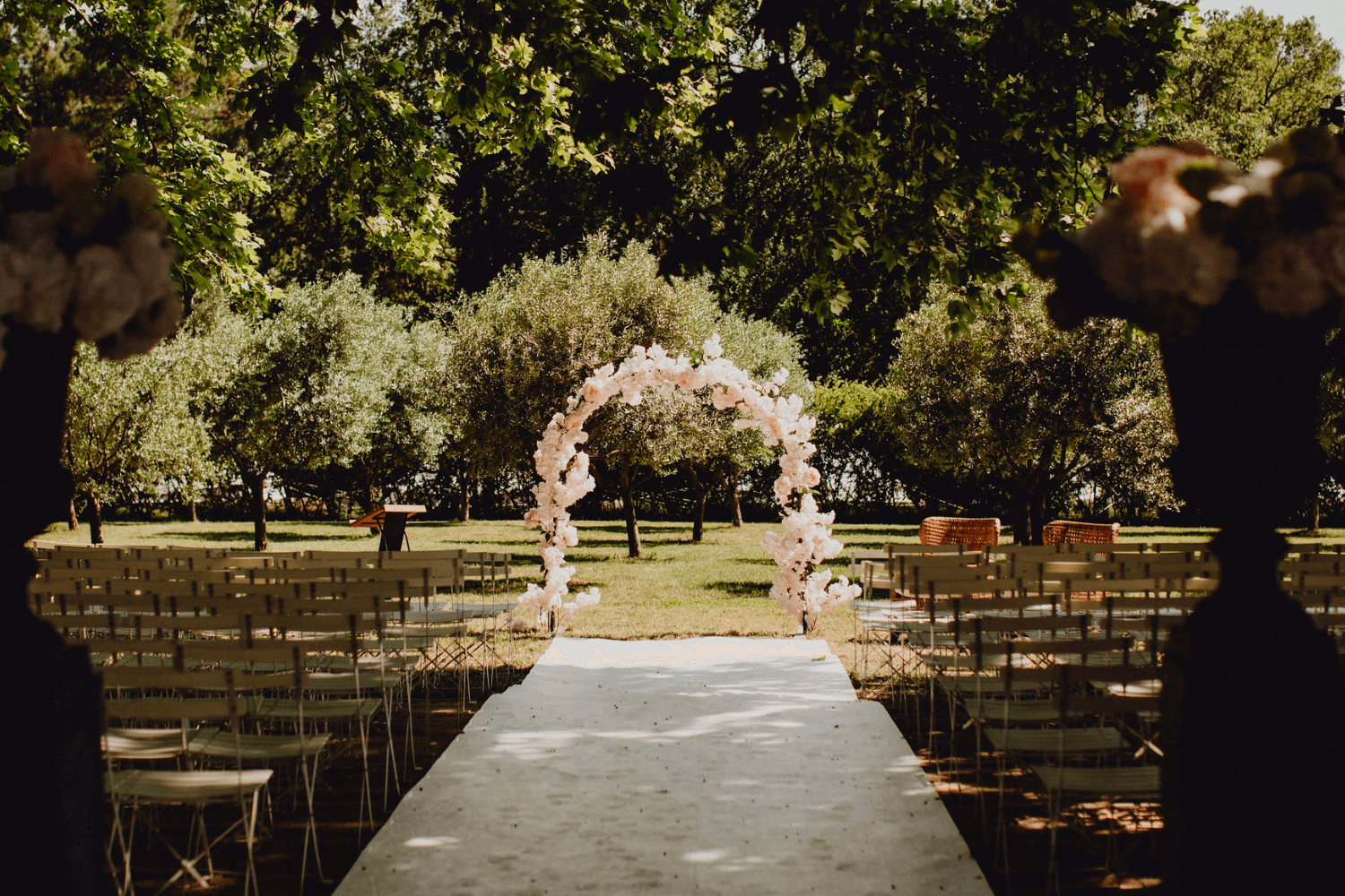 Un lieu de réception de cérémonie laïque extérieur entouré d’arbres, avec une arche de mariage fleurie au centre.