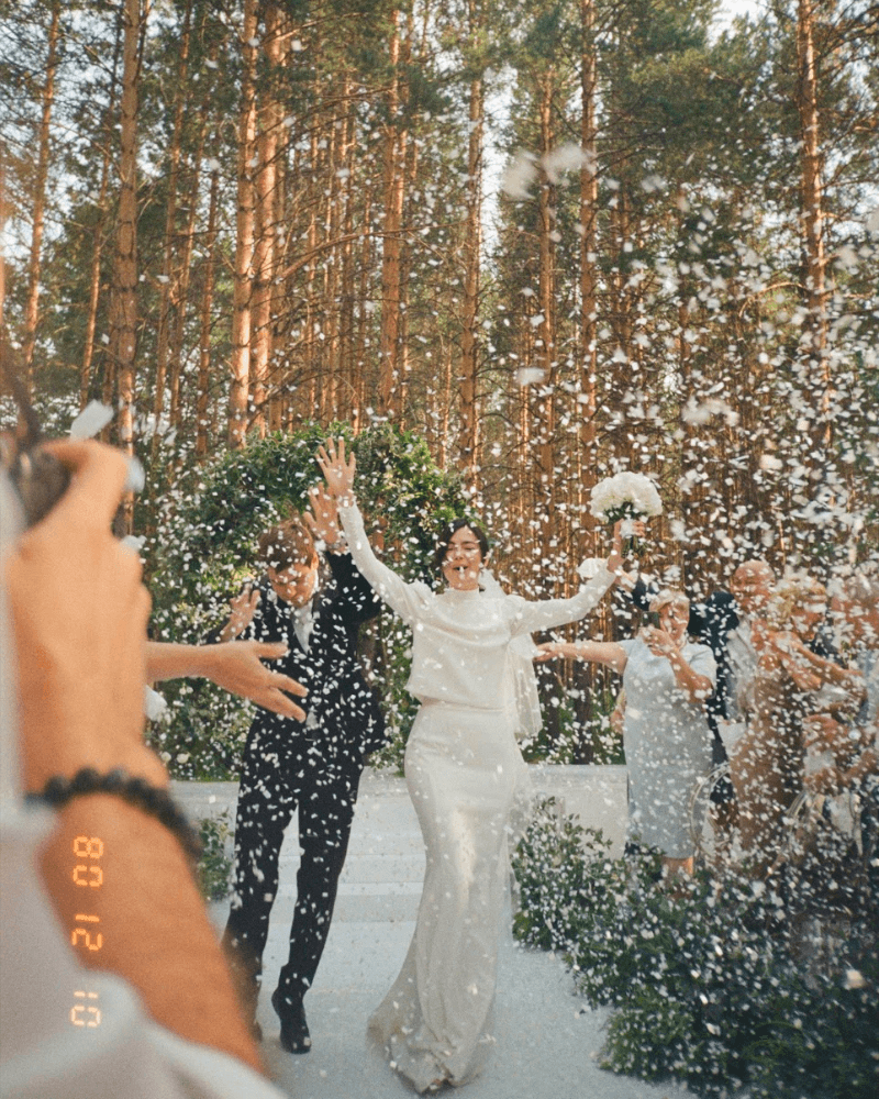 un couple de jeunes mariés à la sortie de leur cérémonie, levant les bras sous les confettis de leurs invités