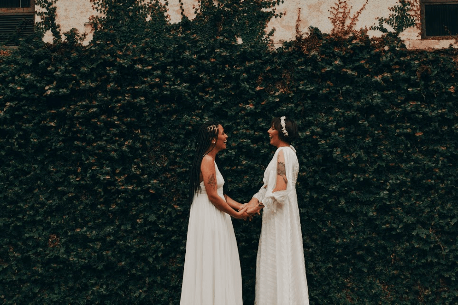 Un couple deux femmes mariées en robes blanches, se faisant face et se tenant les mains en souriant.