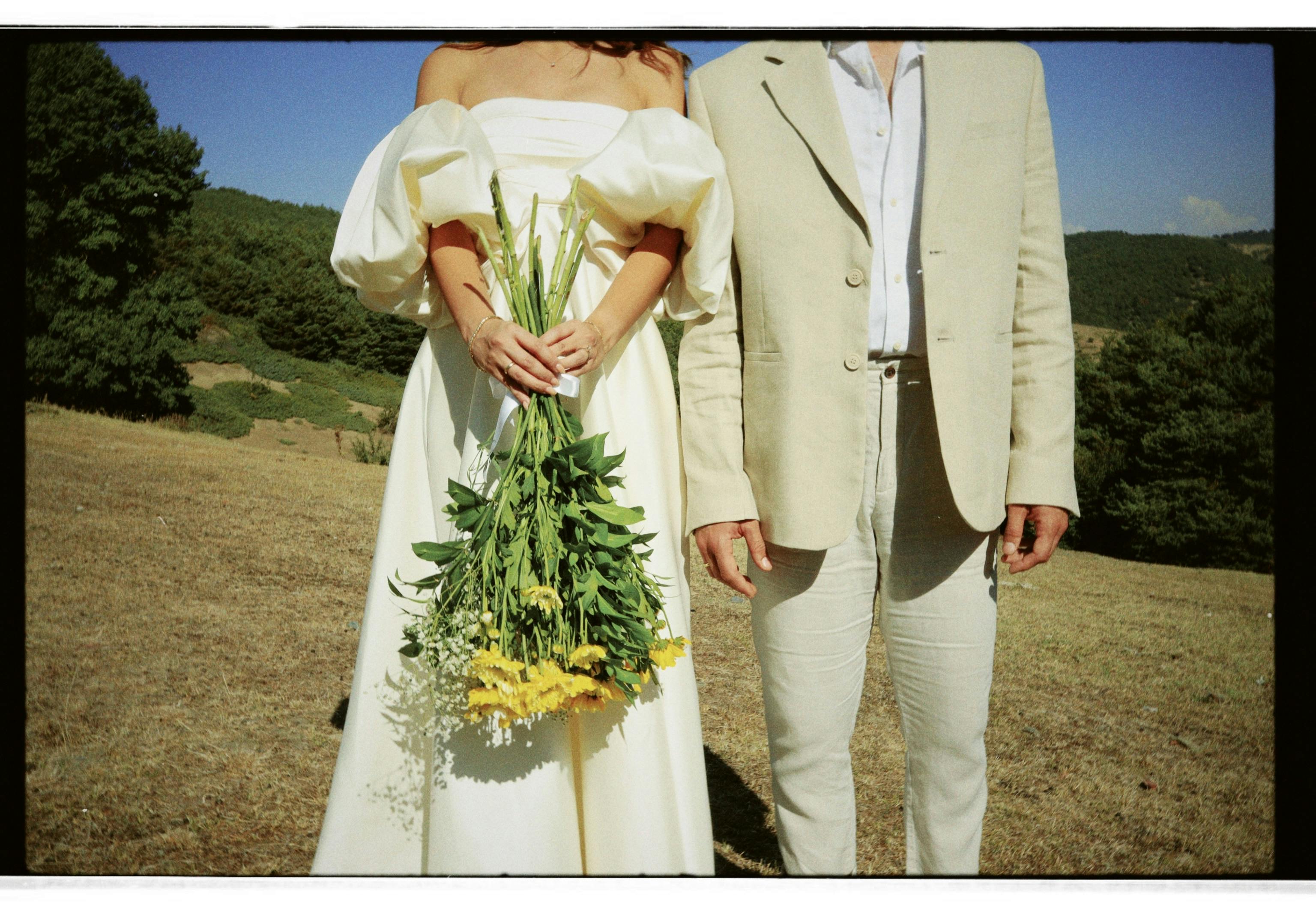 un couple de jeunes mariés, la mariée porte une robe avec manches courtes bouffies et épaules dénudées, le marié porte un costume en lin de couleur beige.