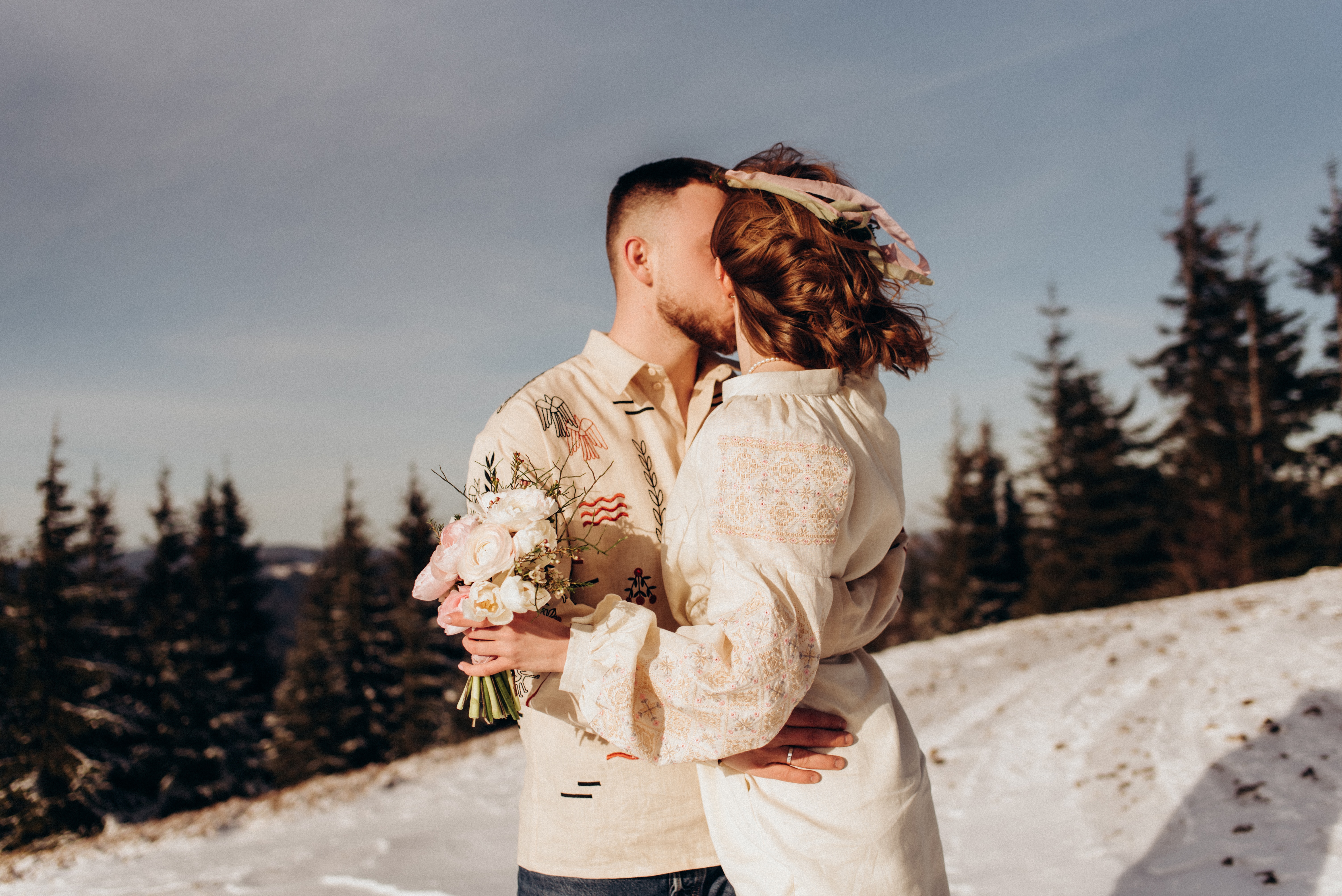 Un couple de jeunes mariés s'embrassant dans la neige. La mariée porte une robe à manches longues et tient un bouquet de fleurs dans une main, le marié porte une chemise.