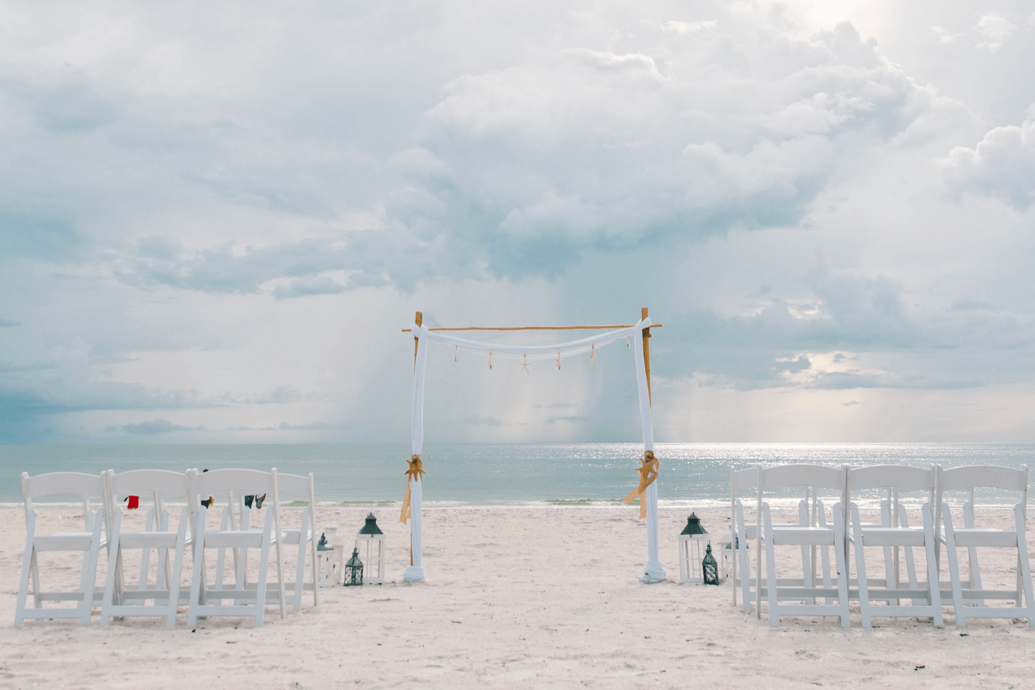 Une cérémonie de mariage sur la plage avec des chaises déposées en rang sur le sable et une arche de mariage agrémentée d’un ruban blanc.