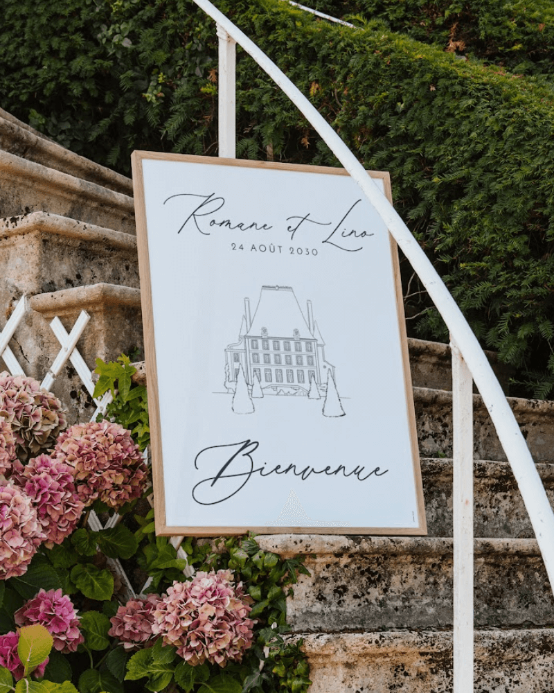 Panneau de bienvenue de mariage avec illustration d’une grande villa en noir et blanc. Le panneau est déposé sur le rebord d’un escalier fleuri.