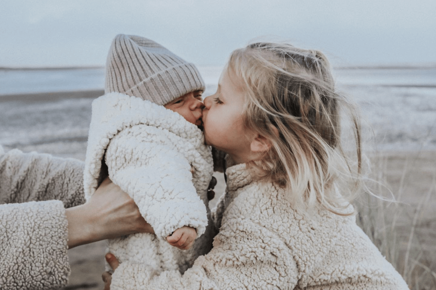 un bébé dans les bras de sa maman, sa grande sœur l’embrassant. Tous deux sont vêtus de tenues d’hiver.