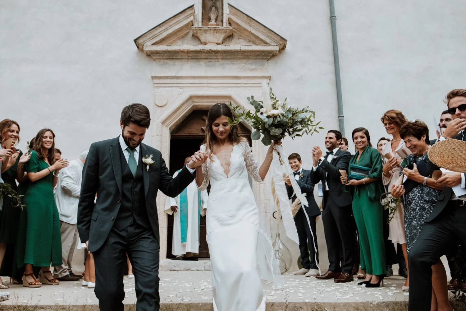 Un couple de jeunes mariés sortant de l’église en se tenant par la main. En arrière plan, on aperçoit les invités applaudissant.