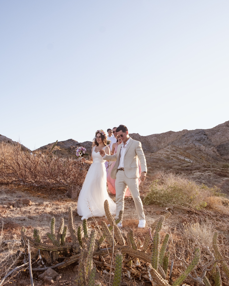 Un couple de jeunes mariés se tenant par la main, dans le désert. Derrière eux, on aperçoit leurs invités.