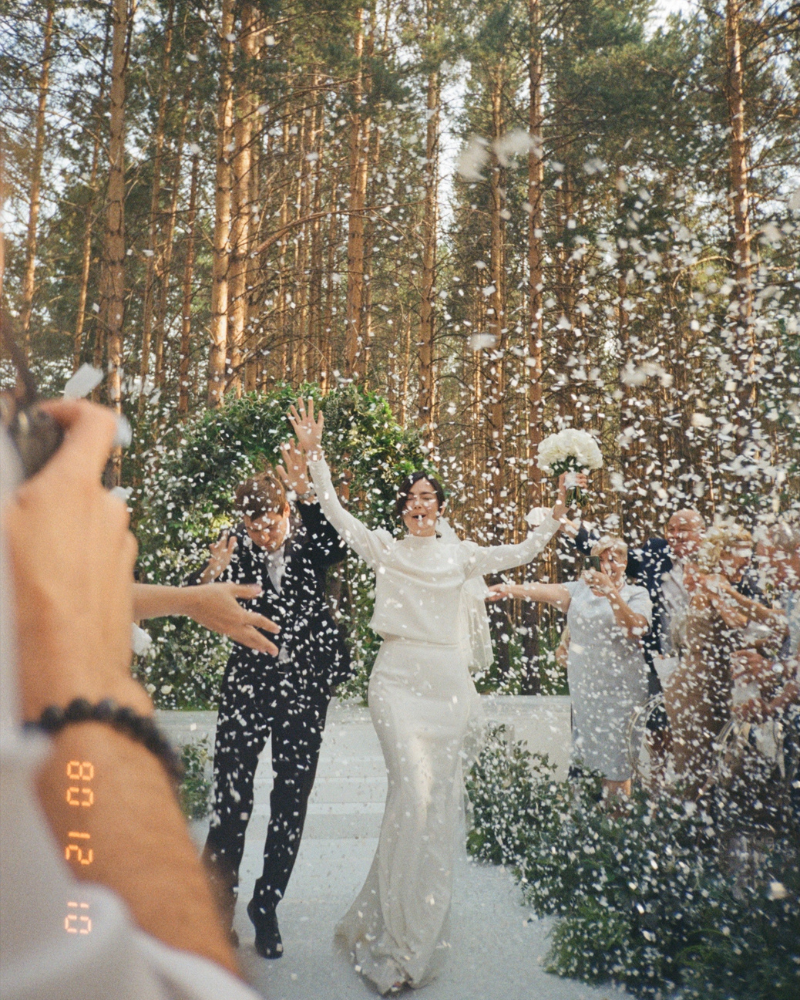 Un couple de jeunes mariés entourés de leurs invités, levant les bras sous une pluie de confettis de neige.