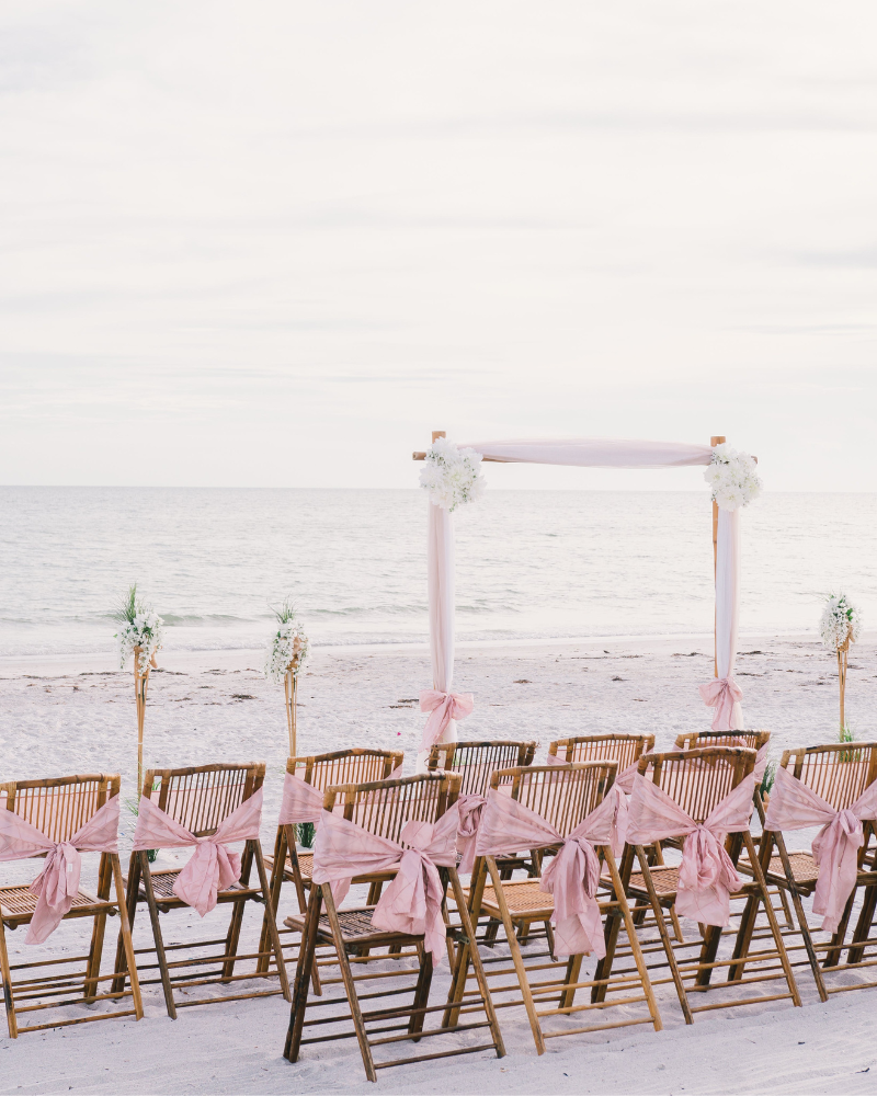 une cérémonie de mariage sur la plage avec des chaises en bois décorées de rubans roses et une arche de mariage décorée de rubans roses et fleurs.