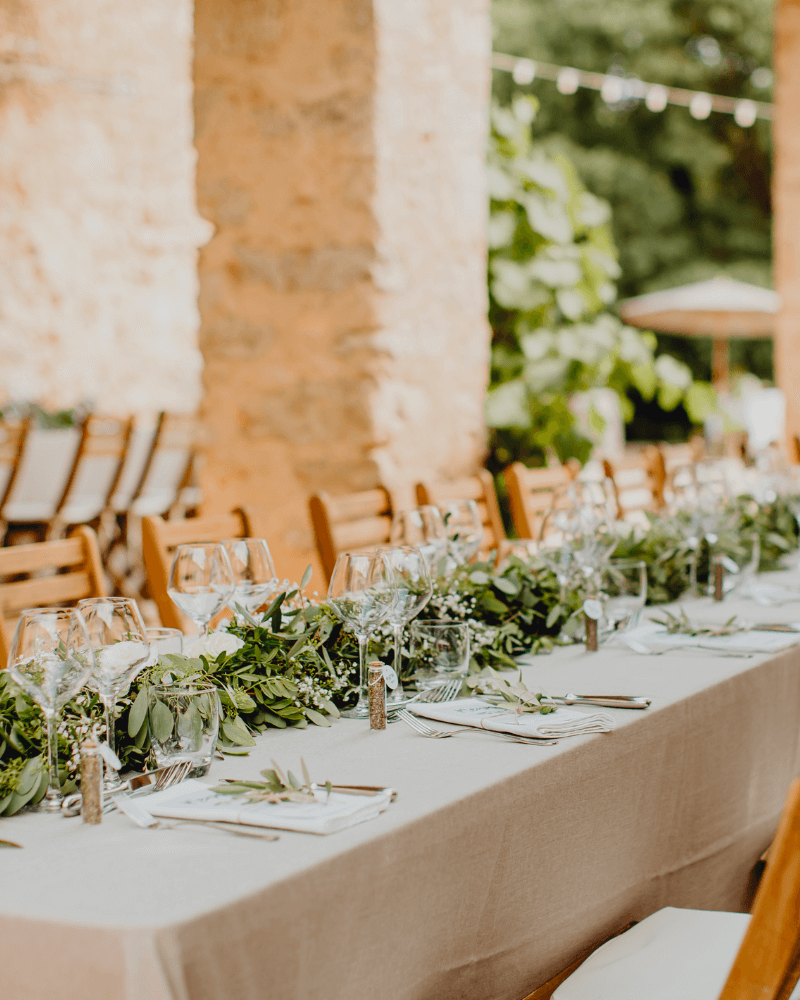 Une table de mariage champêtre recouverte d'une nappe beige et décorée d'un chemin de table végétal.