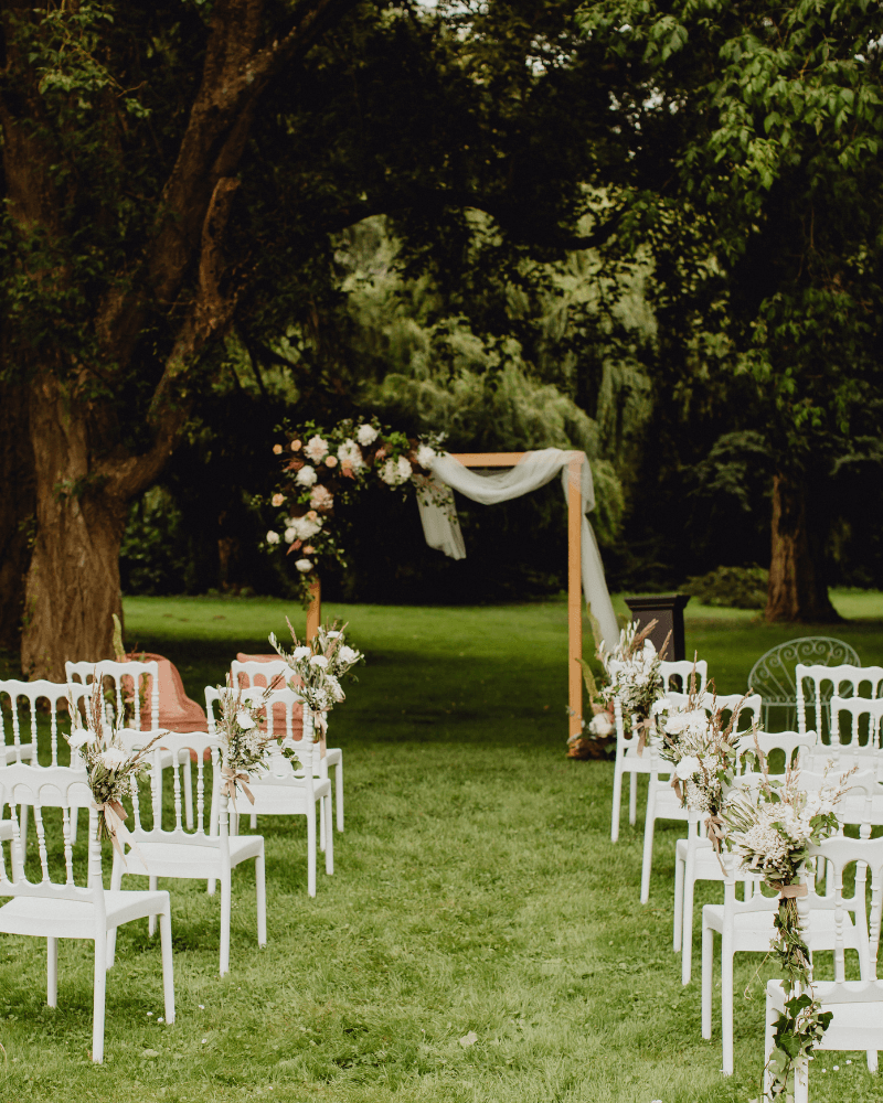 Un lieu de réception de mariage extérieur champêtre avec chaises blanches décorées de fleurs et une arche de mariage en bois fleurie.