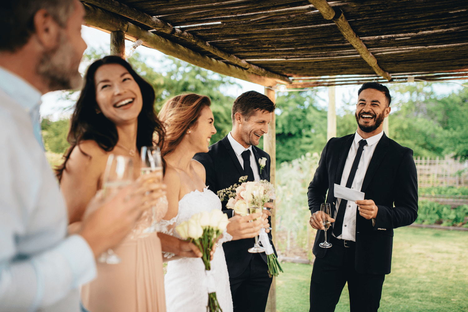 Un témoin de mariage prononçant un discours, tenant dans ses mains un papier et une coupe de champagne. Les mariés et témoins l’écoutent en souriant.