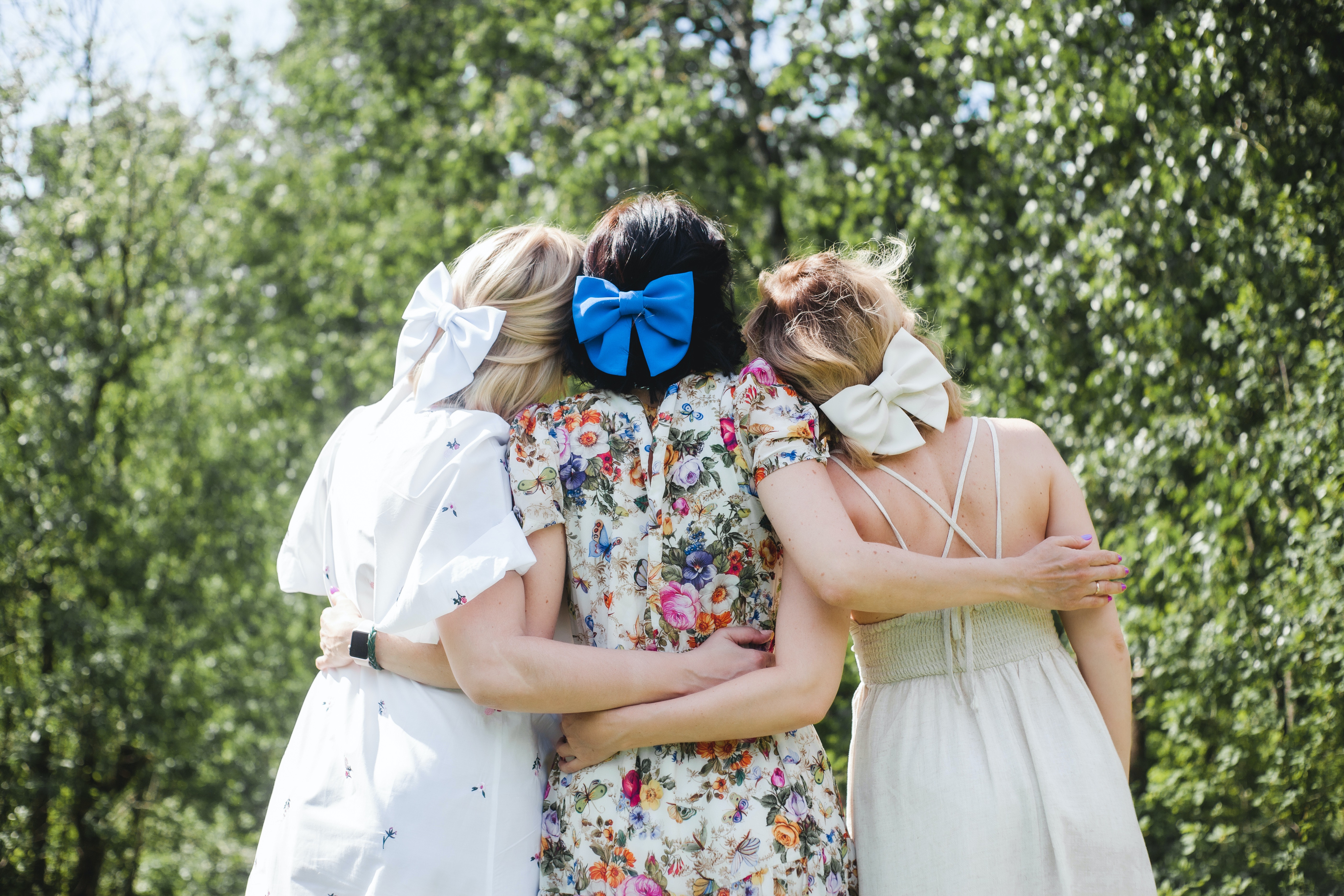 Trois femmes vues de dos, se tiennent dans une accolade. Elles portent un nœud dans leurs cheveux.