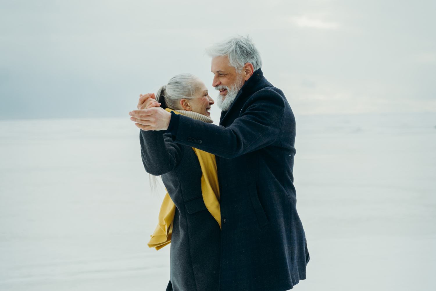 Un couple de mariés seniors aux cheveux blancs, se tenant par la main pour danser. Tous deux sont habillés de noir, la femme a une écharpe jaune autour du cou. Ils sourient.