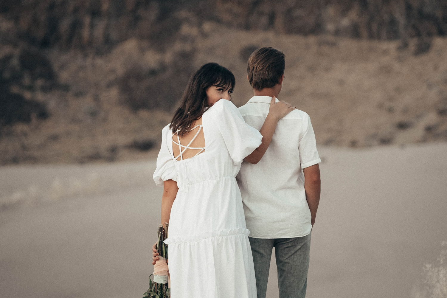 Un couple vêtu de blanc, dos à la caméra. La femme regarde en arrière vers l’objectif en souriant, l’une de ses mains posées sur les épaules de son partenaire.