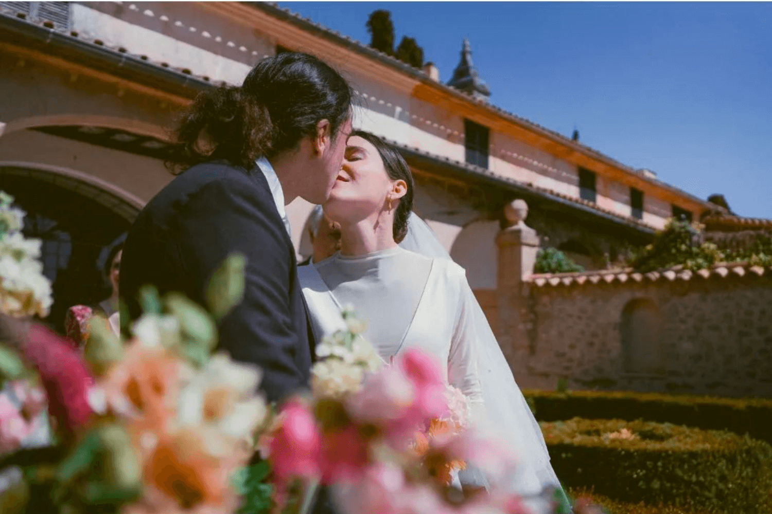 Un couple de jeunes mariés s'embrassant. Au premier plan, on aperçoit un bouquet de fleurs aux tons roses et orangés et, au fond, une grande demeure provençale.