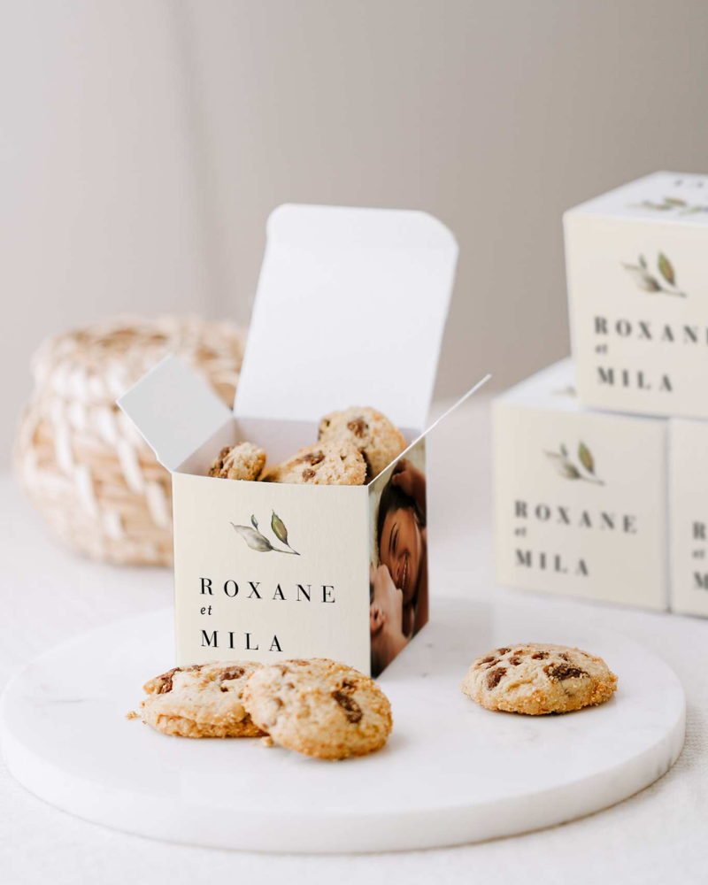 Un petit cube surprise décoré avec les prénoms des mariées, une petite illustration végétale et la photo des mariées. À l'intérieur et autour, des mini cookies.