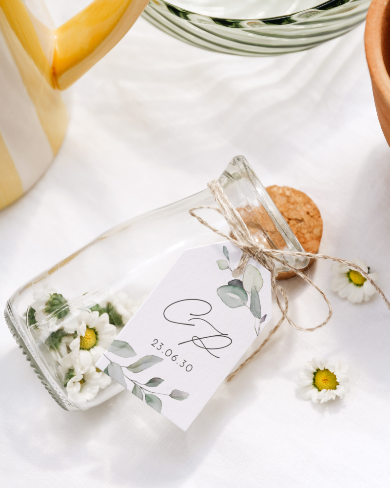 Une petite fiole en verre contenant des marguerites, décorée d'une étiquette personnalisée avec initiales.
