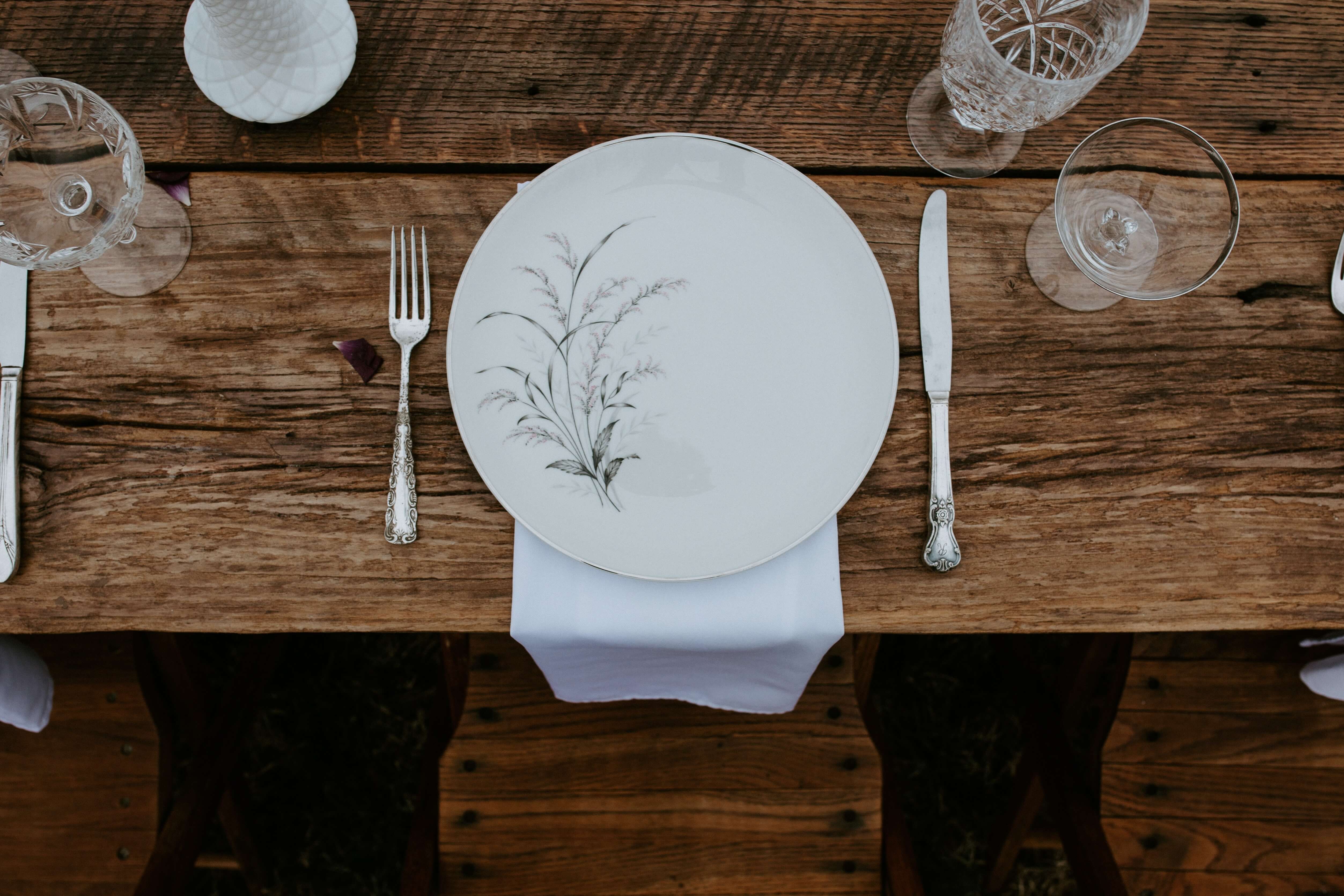Une table en bois dressée, avec une assiette blanche décorée d'épis de blé gris. Autour, couverts et verres.