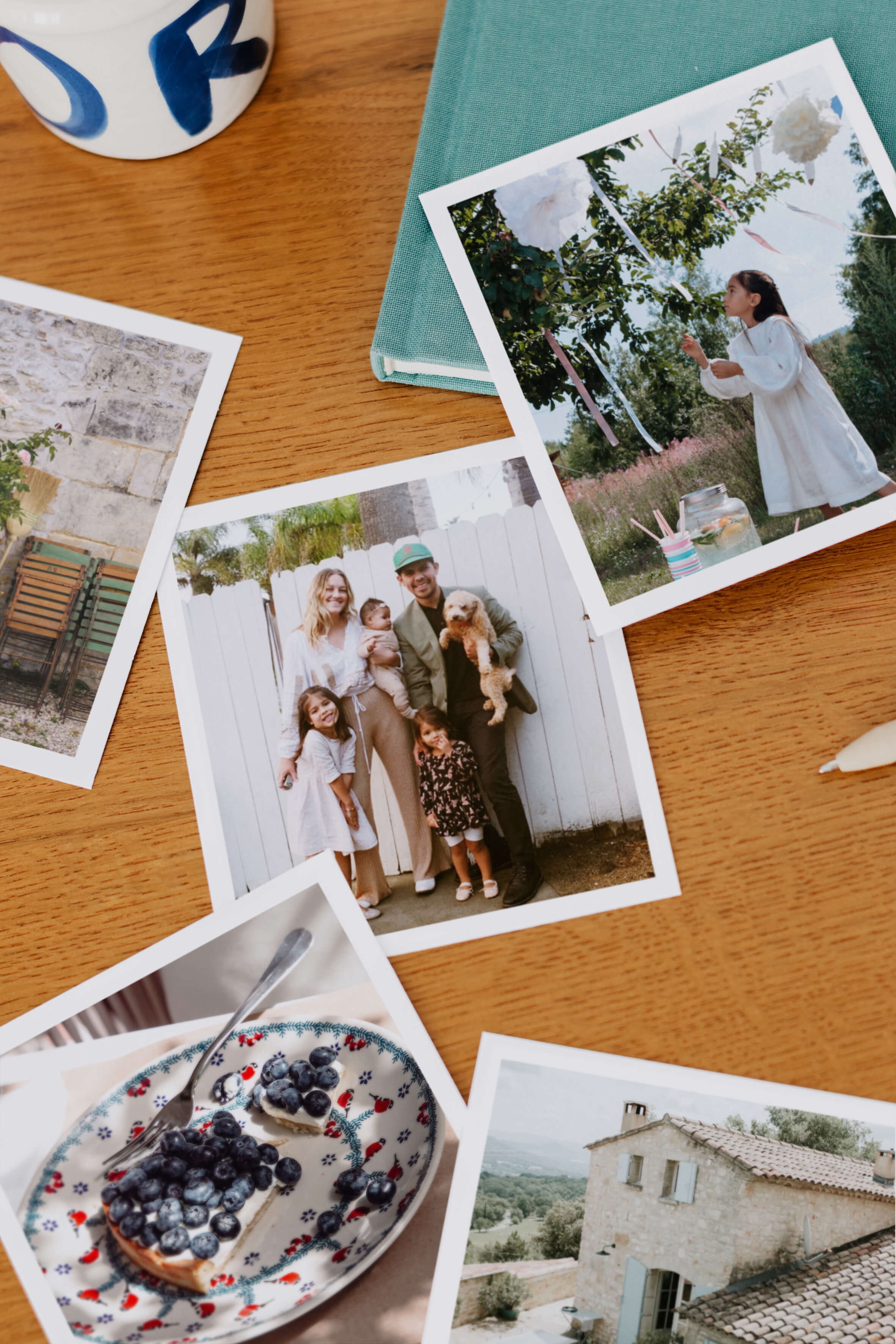 Plusieurs tirages photo polaroid posés sur une table en bois. On aperçoit une famille, une petite fille, une tarte aux myrtilles, une maison de campagne en pierres.