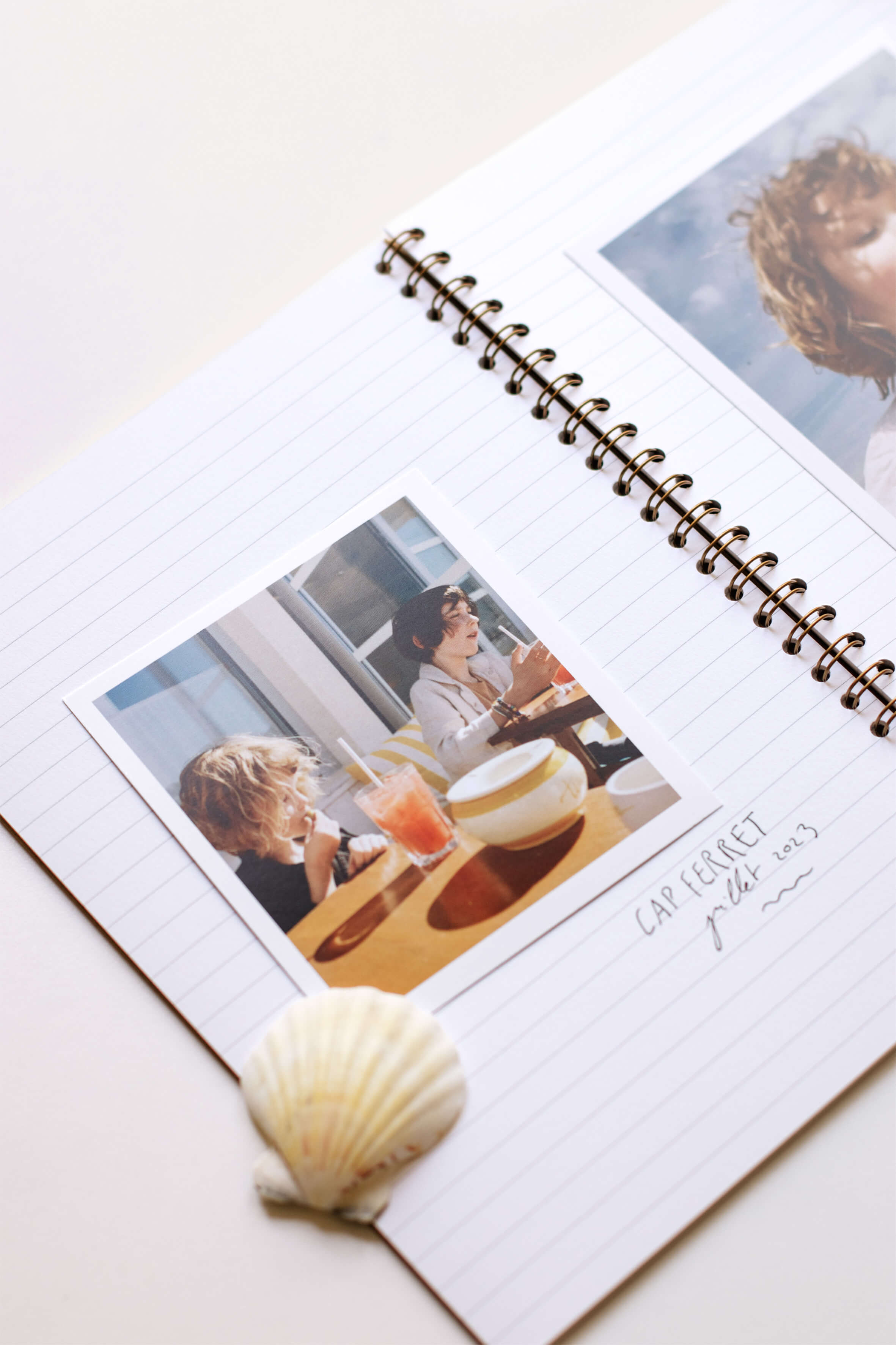 Un tirage photo polaroid collé dans un cahier à spirales avec lignes. Sur la photo, on aperçoit deux enfants à table. La photo est légendée : cap ferret, juillet 2023.