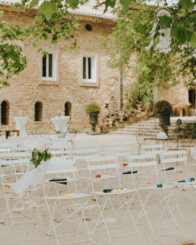 une réception de mariage extérieure : des chaises blanches sont alignées dans une cour de graviers. En arrière plan, on aperçoit une maison de pierres.