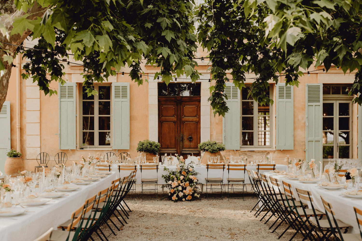 De grandes tables de mariage installées dans une cour extérieure, devant une maison avec des volets peints en vert d’eau.