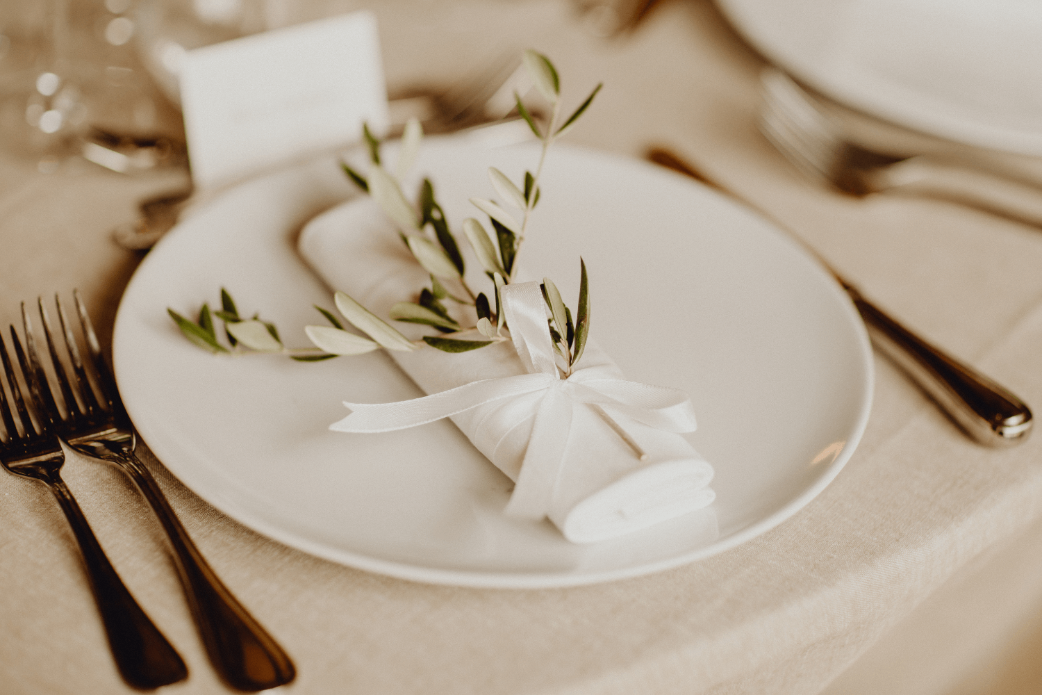 Une assiette blanche entourée de couverts argentés. Au milieu de l'assiette se trouve une serviette blanche, agrémentée d'un ruban blanc et de brins de feuilles.