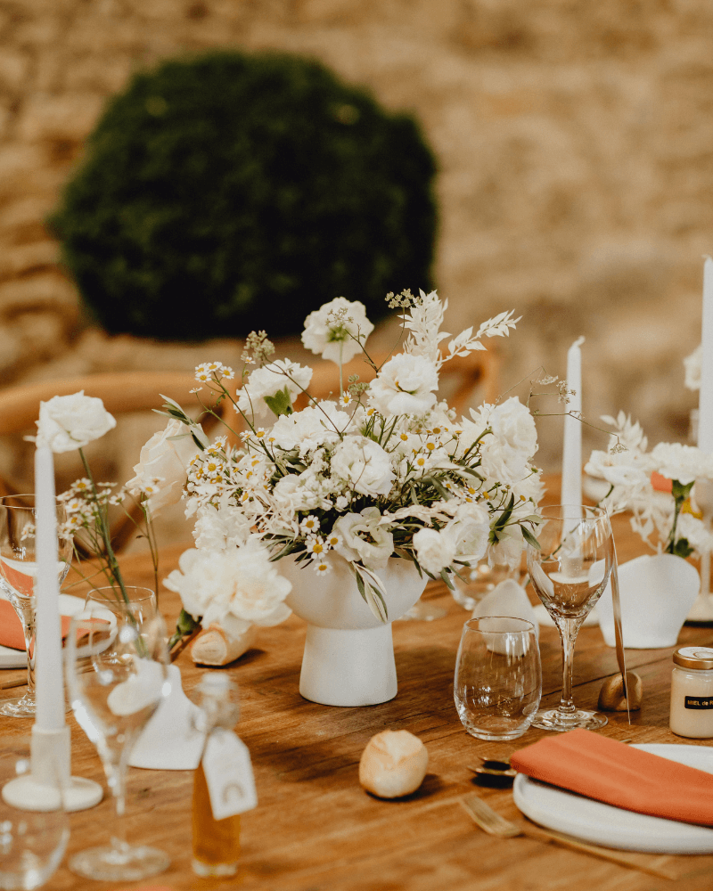 Une table de mariage en bois avec assiettes, verres, couverts, longues bougies et un bouquet de fleurs blanches dans un vase blanc.