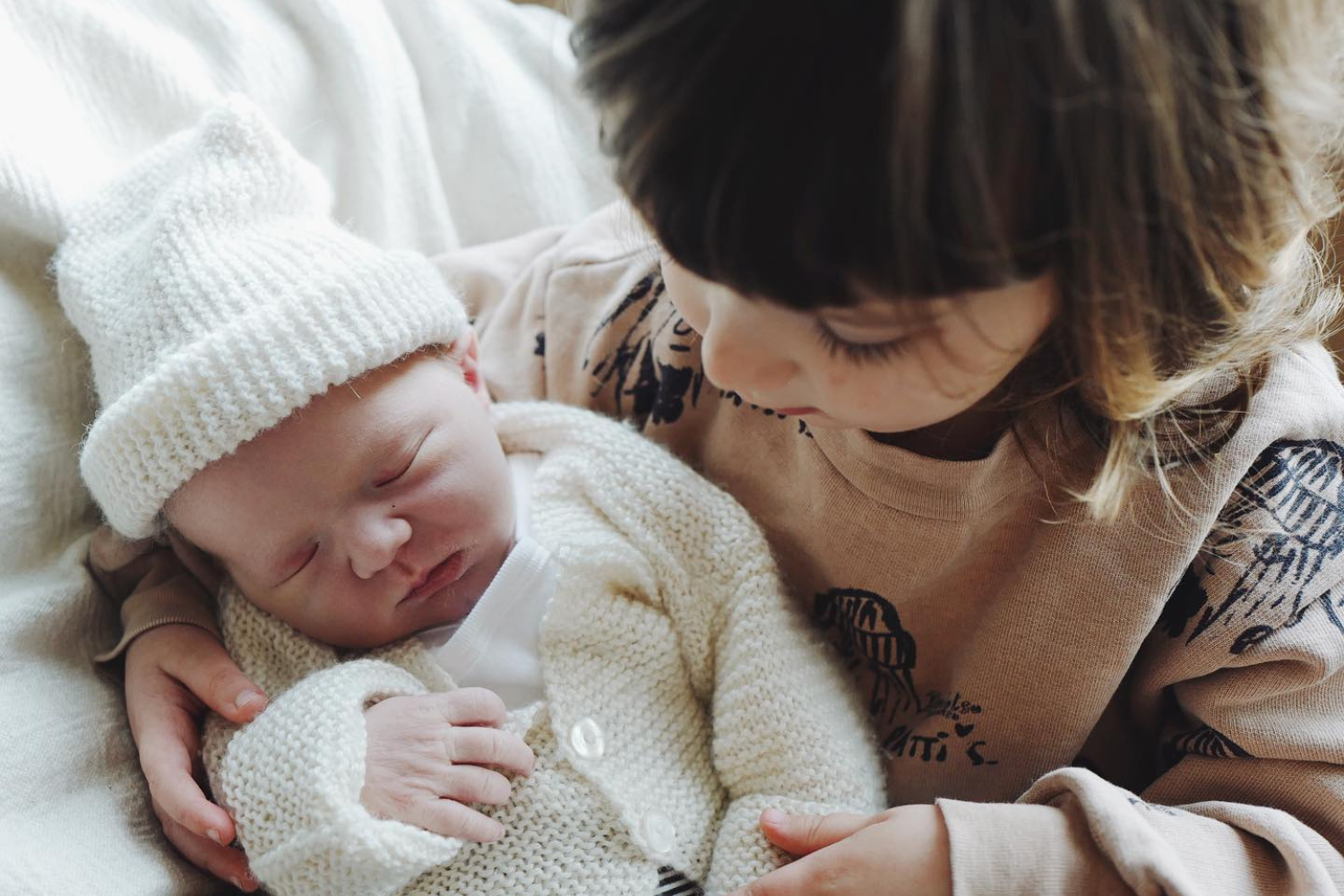 une petite fille tenant dans ses bras un bébé endormi, vêtu d’un gilet et un bonnet de laine blancs.